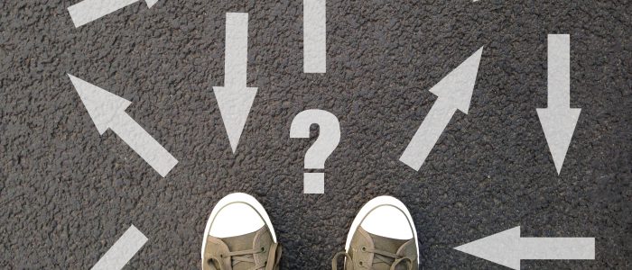 feet standing on asphalt with multitude of arrows in different directions and question mark, confusion choice chaos uncertainty concept